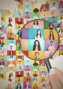 Lupe hebt eine einzelne Person aus einer bunten Übersicht vieler Porträtfotos hervor – Symbolbild für gezielte Personalauswahl und Recruiting.