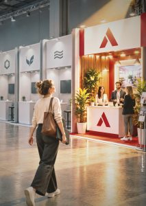Eine Frau geht durch eine moderne Messehalle auf einen hell beleuchteten, rot-weißen Messestand zu, an dem sich drei Personen hinter einem Empfangstresen unterhalten.
