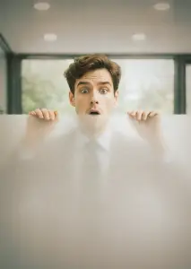 Ein junger Mann mit braunen Haaren schaut überrascht über eine halbtransparente Milchglasscheibe, die er mit beiden Händen festhält. Im Hintergrund ist ein heller, moderner Innenraum mit großen Fenstern zu sehen.