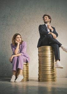 Eine Frau im lilafarbenen Businessanzug sitzt frustriert auf einem niedrigen Stapel goldener Münzen, während ein Mann im dunklen Anzug selbstzufrieden auf einem deutlich höheren Münzstapel sitzt. Die unterschiedlich hohen Stapel symbolisieren den Gender Pay Gap und die ungleiche Bezahlung von Frauen und Männern.