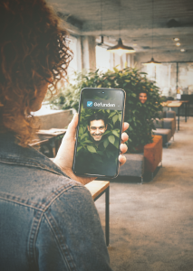 Frau entdeckt versteckten Mann hinter einer Pflanze in modernem Büro mithilfe ihres Smartphones („Gefunden“-Anzeige), Symbol für Social Recruiting und das Finden passiver Talente.