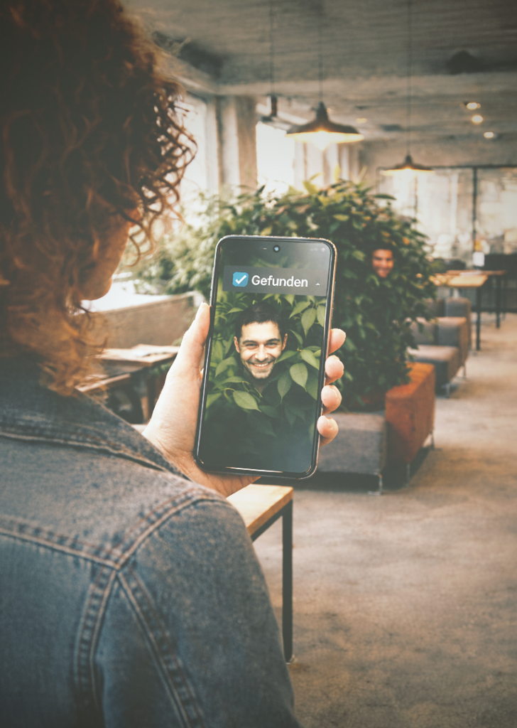 Frau entdeckt versteckten Mann hinter einer Pflanze in modernem Büro mithilfe ihres Smartphones („Gefunden“-Anzeige), Symbol für Social Recruiting und das Finden passiver Talente. KI-generiert